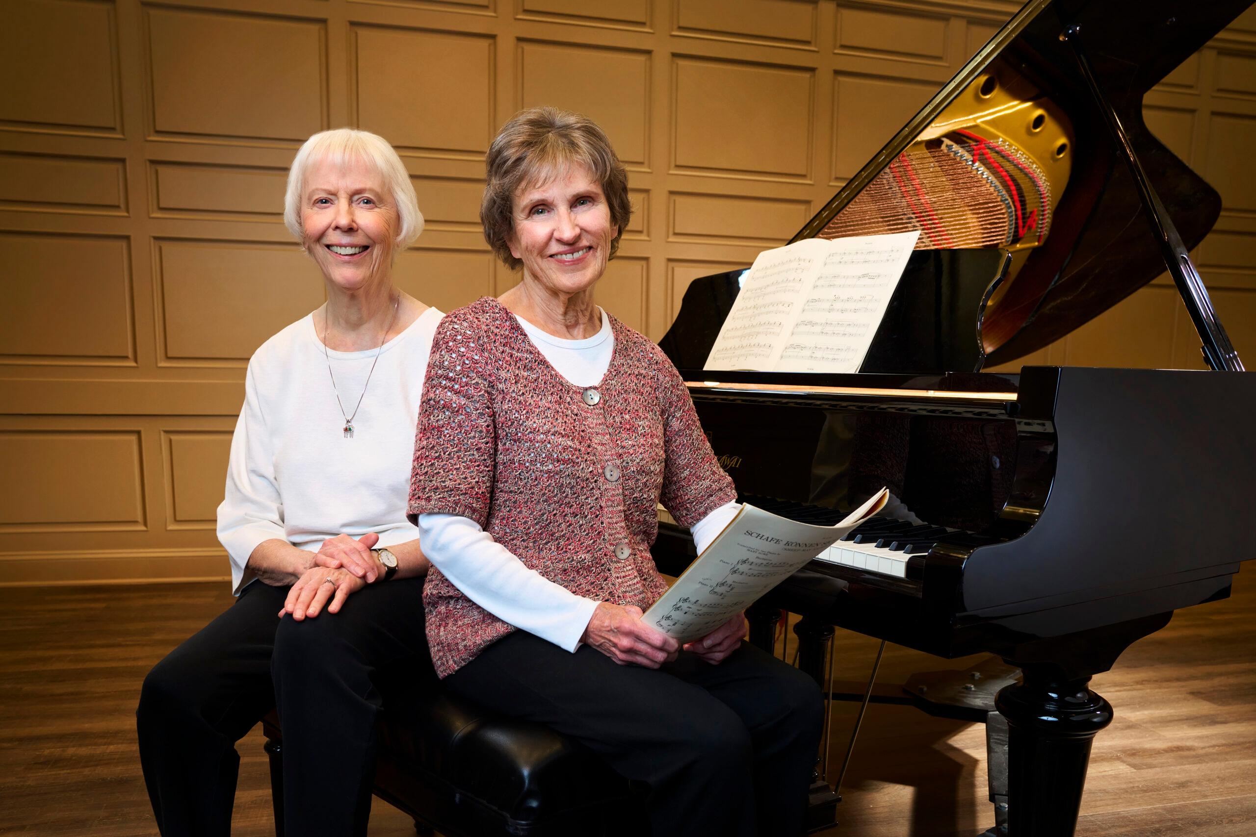 Two pianists sit on the bench of a baby grand piano.