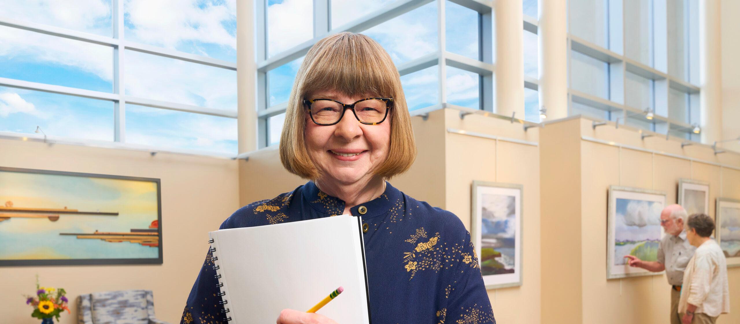 A woman smiles and stands with a notebook in front of several paintings.