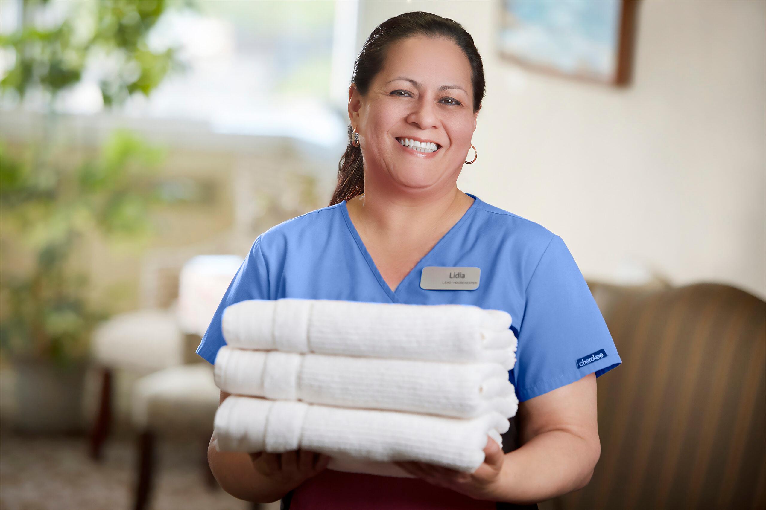 A smiling healthcare employee holds a stack of towels.